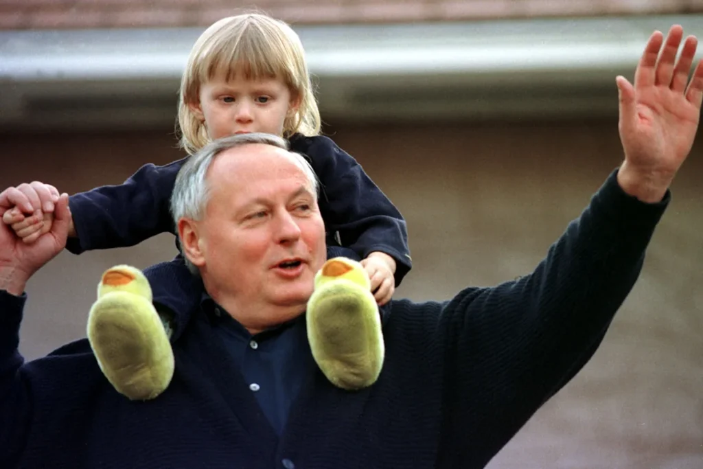 Oskar Lafontaine trägt seinen kleinen Sohn Carl-Maurice auf den Schultern und winkt mit erhobenem Arm.