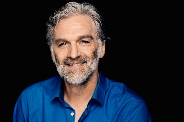 Schauspieler Walter Sittler mit grauem Bart und blauem Hemd lächelt vor schwarzem Hintergrund